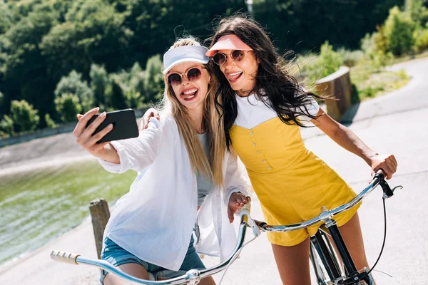 Enfoque selectivo de chicas rubias y morenas mostrando lenguas mientras toma selfie en bicicletas - foto de stock