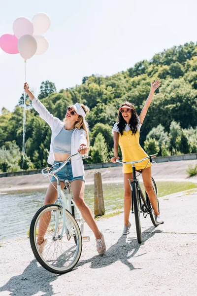 Happy blonde and brunette friends riding bikes with balloons near river in summer — Stock Photo