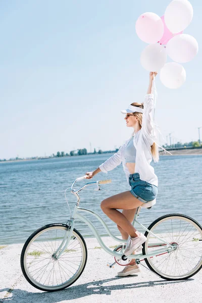Vista lateral de sorrir menina loira andar de bicicleta com balões perto do rio no verão — Fotografia de Stock