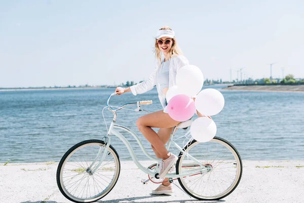 Vista lateral da menina loira feliz andar de bicicleta com balões perto do rio no verão — Fotografia de Stock