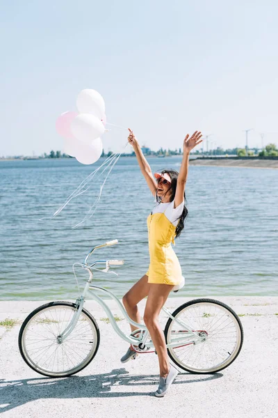 Vista lateral de la chica morena feliz montar en bicicleta con globos y manos en el aire cerca del río en verano - foto de stock