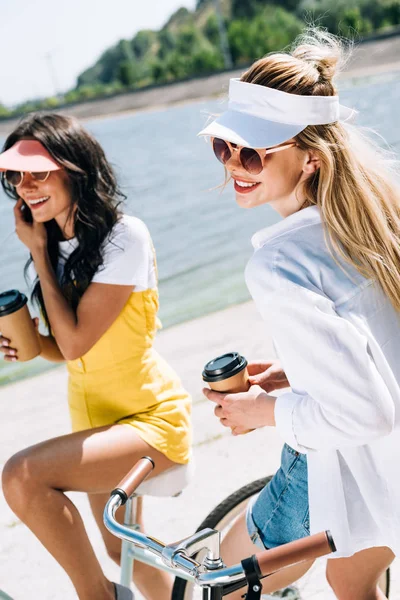 Happy blonde and brunette friends on bikes with coffee to go near river in summer — Stock Photo