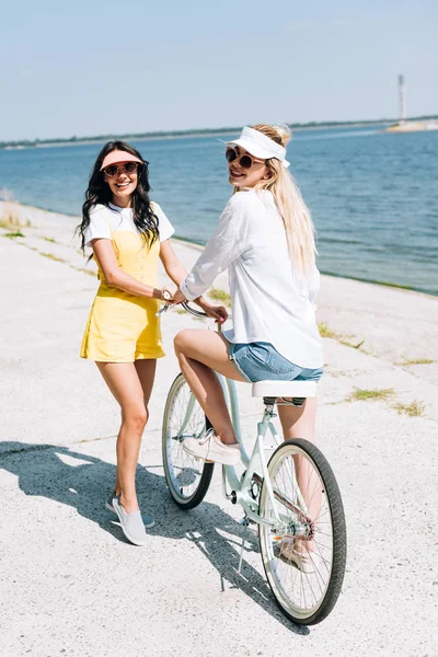 Happy blonde and brunette friends with bike near river in summer — Stock Photo