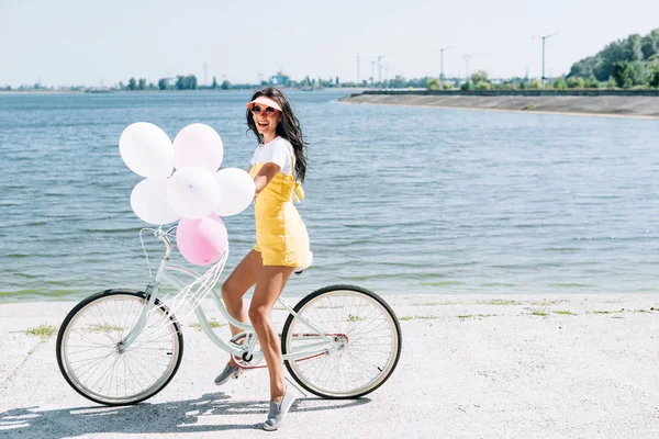 Vista lateral da bela morena menina andar de bicicleta com balões perto do rio — Fotografia de Stock
