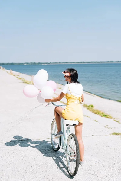 Back view of beautiful brunette girl riding bike with balloons near river — Stock Photo