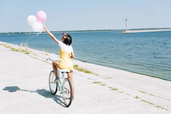 Вид сзади брюнетка девушка на велосипеде с воздушными шарами возле реки — стоковое фото