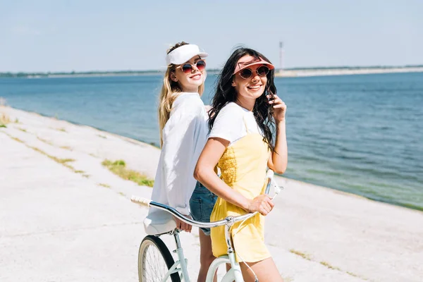 Sorridente bionda e bruna ragazze con bici vicino al fiume in estate — Foto stock