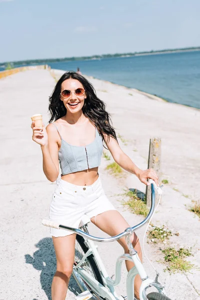 Happy brunette girl with ice cream riding bike near river in summer — Stock Photo
