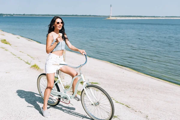 Smiling brunette girl with ice cream riding bike near river in summer — Stock Photo