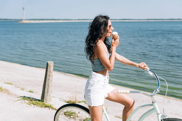 Side view of brunette girl eating ice cream and riding bike near river in summer — Stock Photo