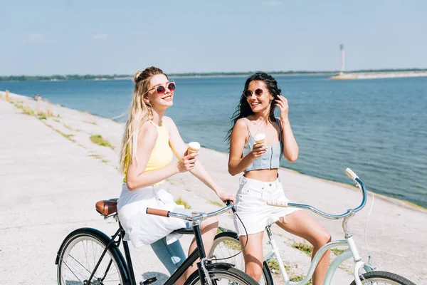 Chicas rubias y morenas felices montando bicicletas con helado cerca del río en verano - foto de stock