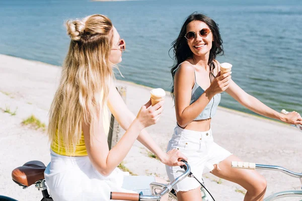 Smiling blonde and brunette girls riding bikes with ice cream near river in summer — Stock Photo