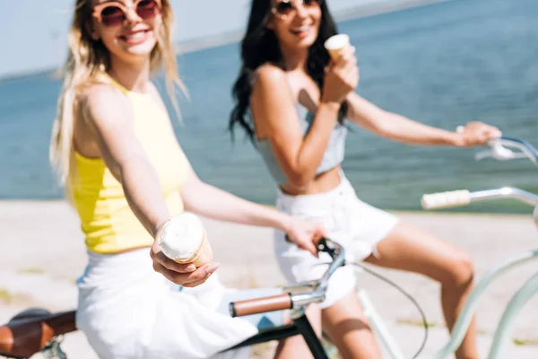 Selective focus of happy blonde girl showing ice cream while riding bike with friend — Stock Photo