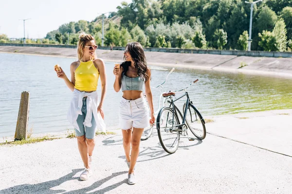 Sonrientes chicas rubias y morenas cerca de bicicletas con helado en el río en verano - foto de stock