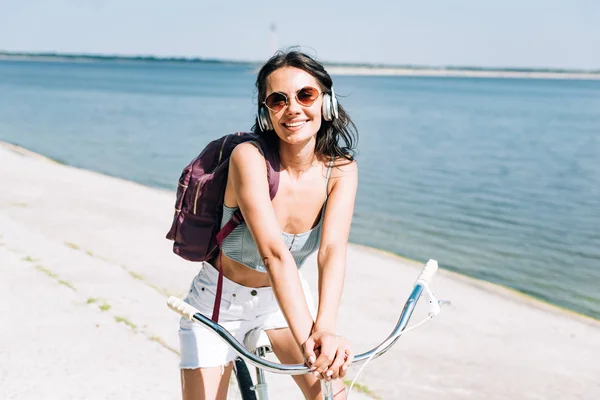 Smiling girl with backpack biking and listening music in headphones near river in summer — Stock Photo