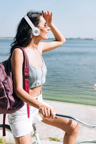 Chica morena feliz con mochila en bicicleta y escuchar música en los auriculares cerca del río - foto de stock