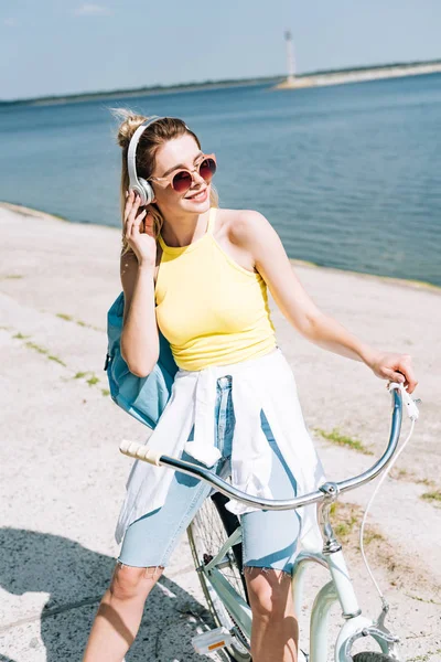 Ragazza bionda con zaino in bicicletta e ascoltare musica in cuffie vicino al fiume in estate — Foto stock