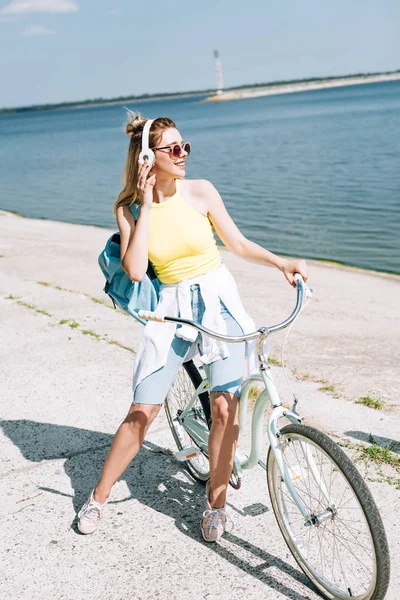 Chica rubia en bicicleta y escuchar música en los auriculares cerca del río en verano - foto de stock