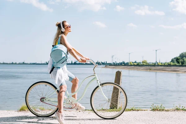 Вид сбоку девушки с рюкзаком, катающейся на велосипеде и слушающей музыку в наушниках у реки летом — стоковое фото