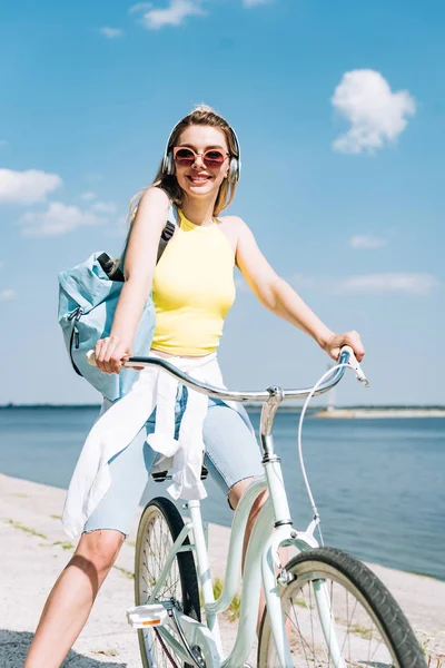 Chica con mochila en bicicleta y escuchar música en los auriculares cerca del río - foto de stock