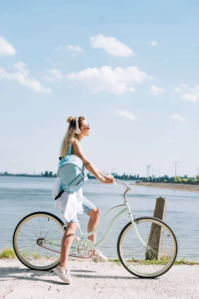 Вид сбоку девушки с рюкзаком, катающейся на велосипеде в наушниках у реки летом — стоковое фото