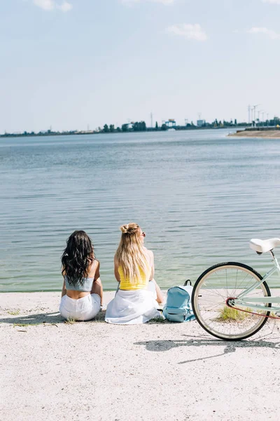 Visão traseira de meninas loiras e morenas sentadas perto de bicicleta e rio no verão — Fotografia de Stock