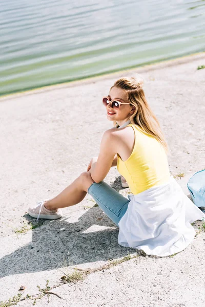 Blonde happy girl in sunglasses with backpack sitting near river in summer — Stock Photo