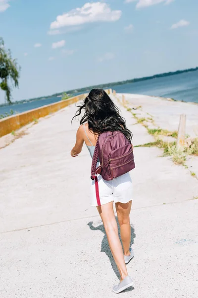 Вид ззаду брюнетка дівчина гуляє з рюкзаком біля річки влітку — стокове фото