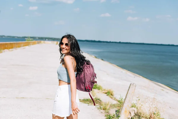 Chica morena feliz caminando con la mochila cerca del río en verano - foto de stock