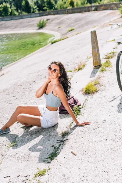 Hermosa chica morena con bicicleta sentado cerca del río en verano - foto de stock