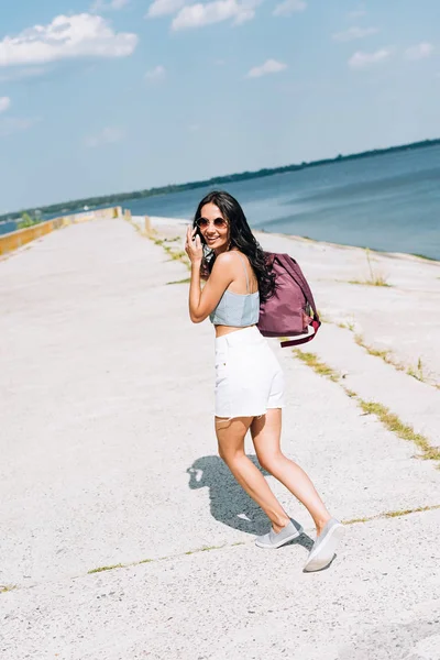 Chica morena feliz en gafas de sol caminando con mochila cerca del río en verano - foto de stock