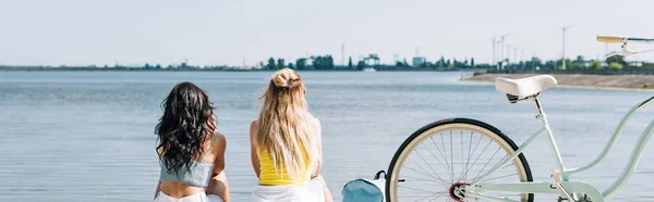 Back view of blonde and brunette friends sitting near bike and river in summer, panoramic shot — Stock Photo