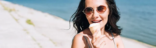 Plano panorámico de chica morena comiendo helado cerca del río en verano - foto de stock