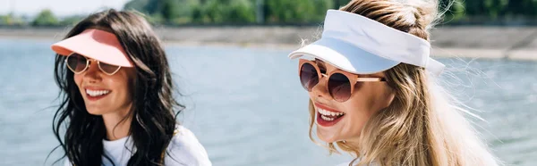 Panoramic shot of happy blonde and brunette girls in sunglasses near river — Stock Photo