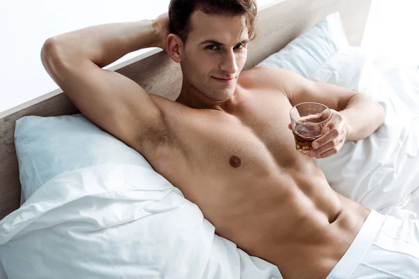 Young sexy man lying on bed and holding glass with whiskey — Stock Photo