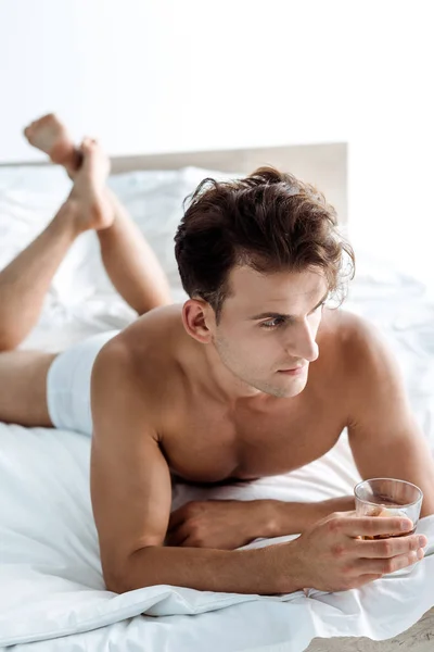 Selective focus of shirtless man lying on bed and holding glass with whiskey — Stock Photo