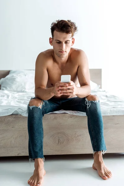 Hombre guapo en jeans usando teléfono inteligente en el dormitorio - foto de stock