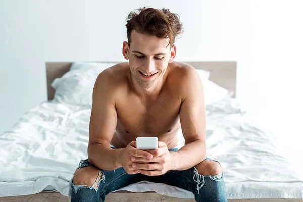 Cheerful man in jeans using smartphone in bedroom — Stock Photo