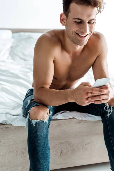 Щасливий чоловік в джинсах, використовуючи смартфон у спальні — стокове фото