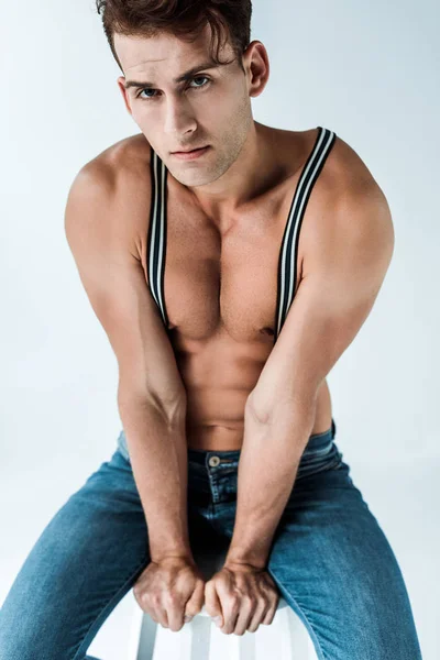 Sexy man in suspenders sitting on chair and looking at camera on white — Stock Photo