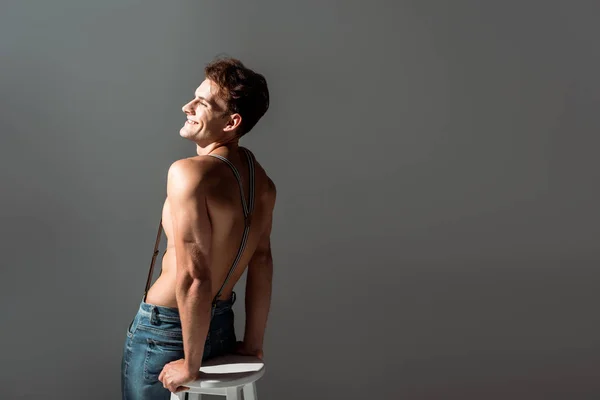 Feliz hombre sin camisa en tirantes cerca de silla blanca aislado en gris - foto de stock