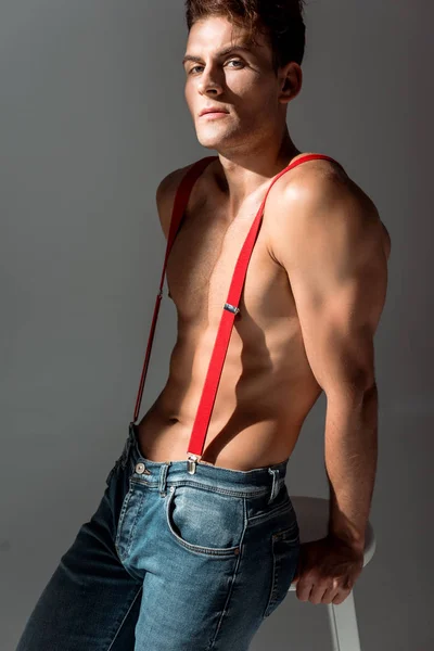 Shirtless man in red suspenders looking at camera and standing near white chair isolated on grey — Stock Photo