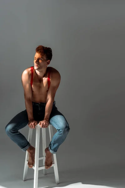 Мускулистый мужчина в красных подтяжках сидит на стуле на сером — стоковое фото