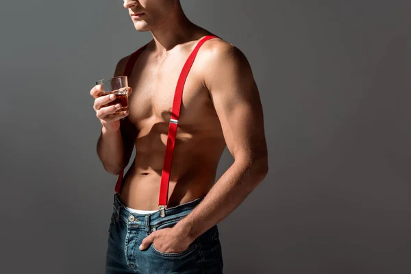 Обрезанный вид человека без рубашки, держащего стакан виски и стоящего с рукой в кармане, изолированного на сером — стоковое фото