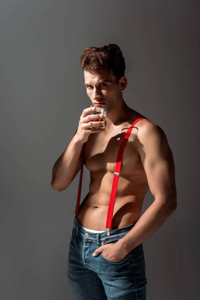 Shirtless man holding glass of whiskey and standing with hand in pocket isolated on grey — Stock Photo