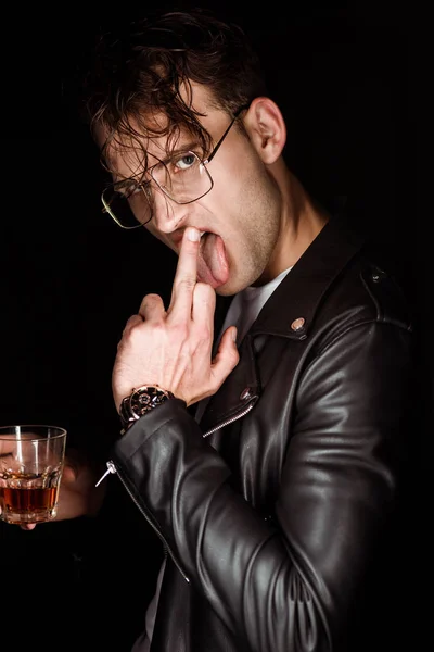 Brutaler Mann zeigt Mittelfinger, streckt Zunge heraus und hält Glas Whiskey isoliert auf schwarz — Stockfoto