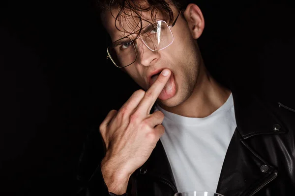 Brutal man in glasses showing middle finger while sticking out tongue isolated on black — Stock Photo