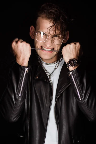 Hombre emocional en gafas mordiendo cadenas metálicas aisladas en negro - foto de stock