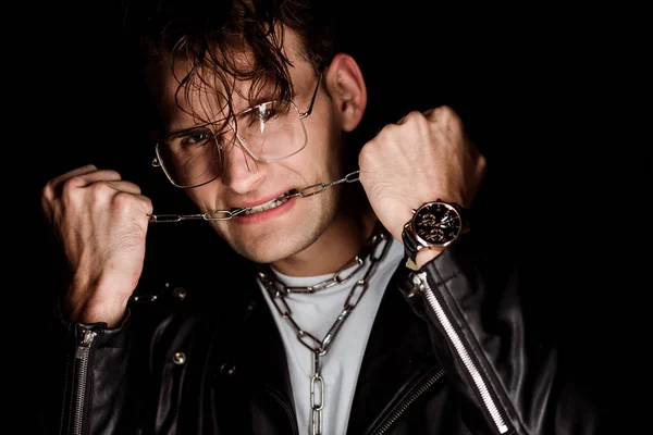 Brutal hombre en gafas mordiendo cadenas metálicas aisladas en negro - foto de stock