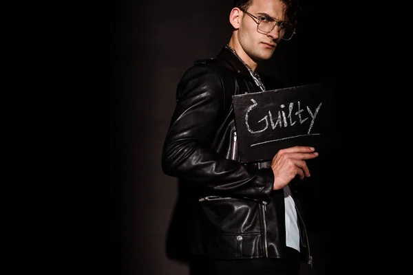 Uomo alla moda in bicchieri tenendo bordo gesso con iscrizione colpevole su nero — Foto stock